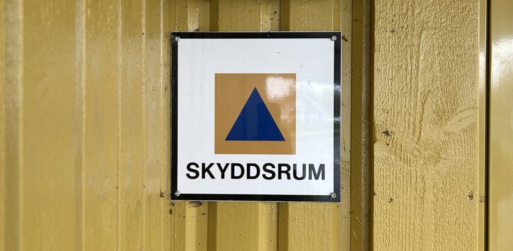 Gul träfasad med en skylt som indikerar skyddsrum, orange fyrkant med blå triangel och texten skyddsrum.